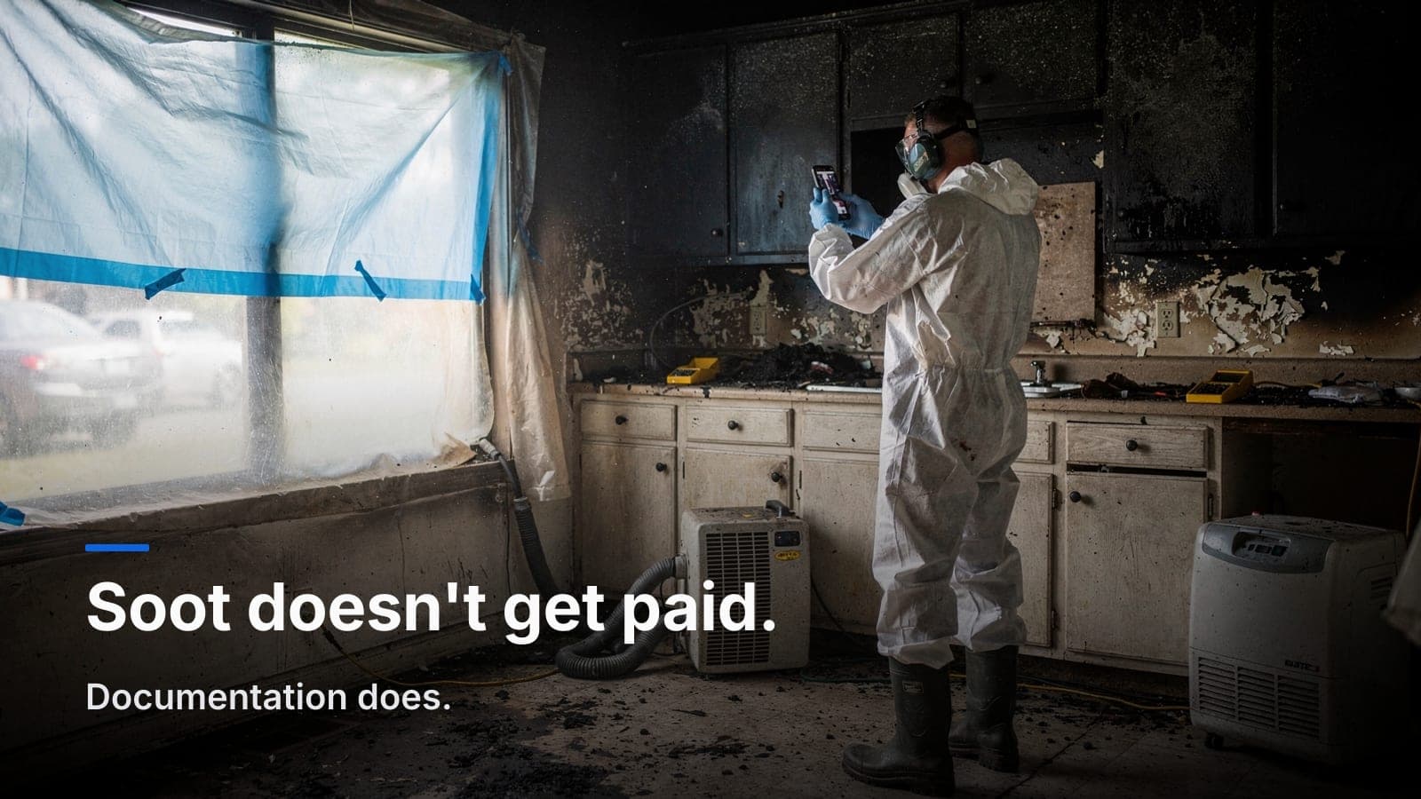 Technician in a soot-stained Tyvek suit and respirator photographing blackened kitchen cabinets, light coming through a tarped window.