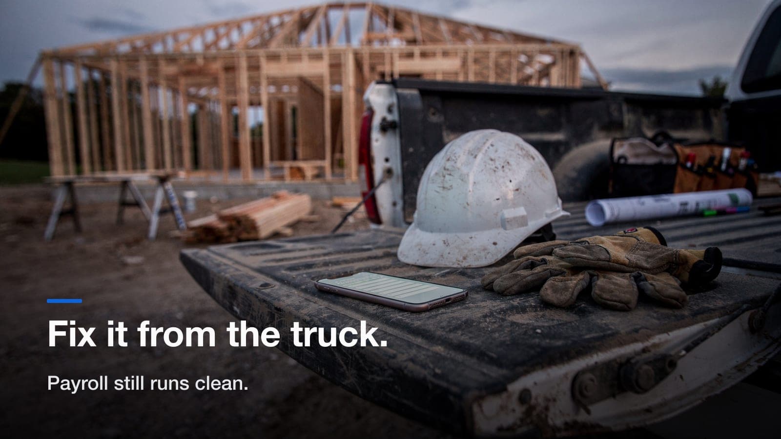 A phone, a hard hat and work gloves on a pickup tailgate at dusk with a framed house behind.